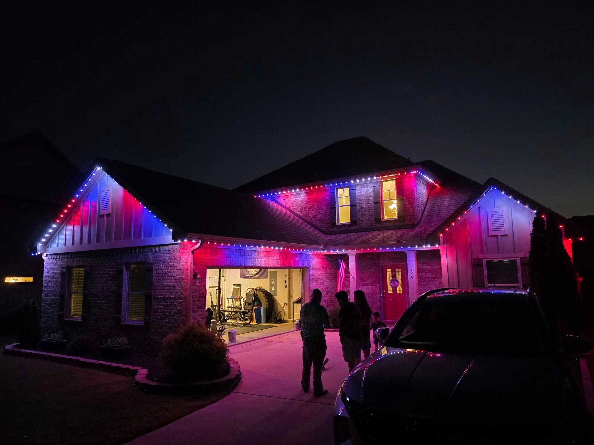 House decorated with red, white, and blue lights; people standing in front of the garage; dark car in the foreground.