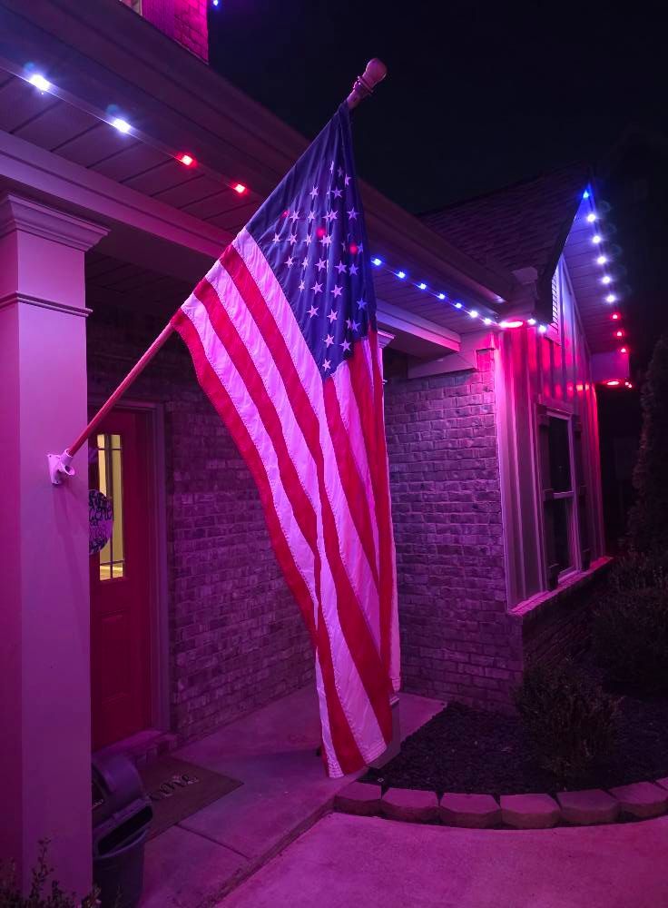 American flag hanging on a house, illuminated with red and blue lights at night.