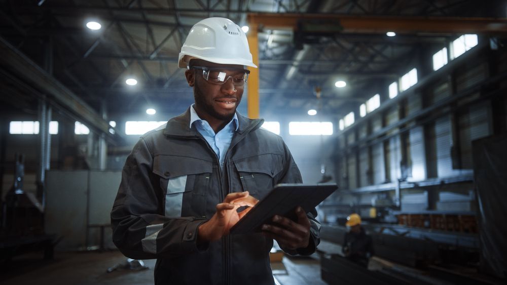 Man in hard hat and safety glasses using a tablet in a factory.