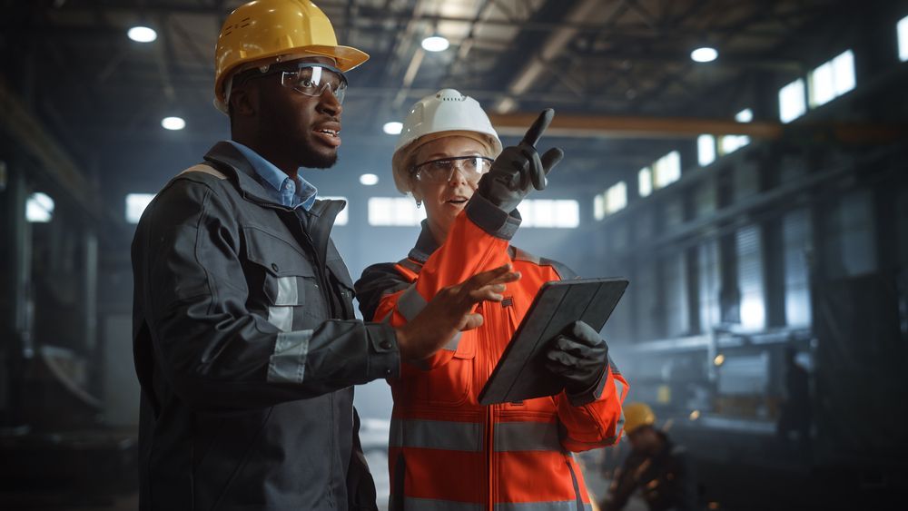 Two factory workers in safety gear, one pointing, looking at a tablet.