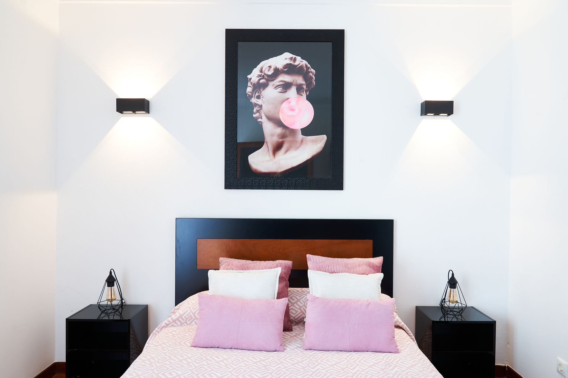 Bedroom with a bed, nightstands, wall art of a statue blowing bubble gum, and two wall-mounted lights.