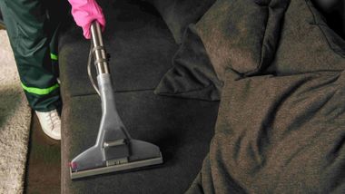 Person using a vacuum cleaner on a dark carpet beside a couch, wearing pink gloves.