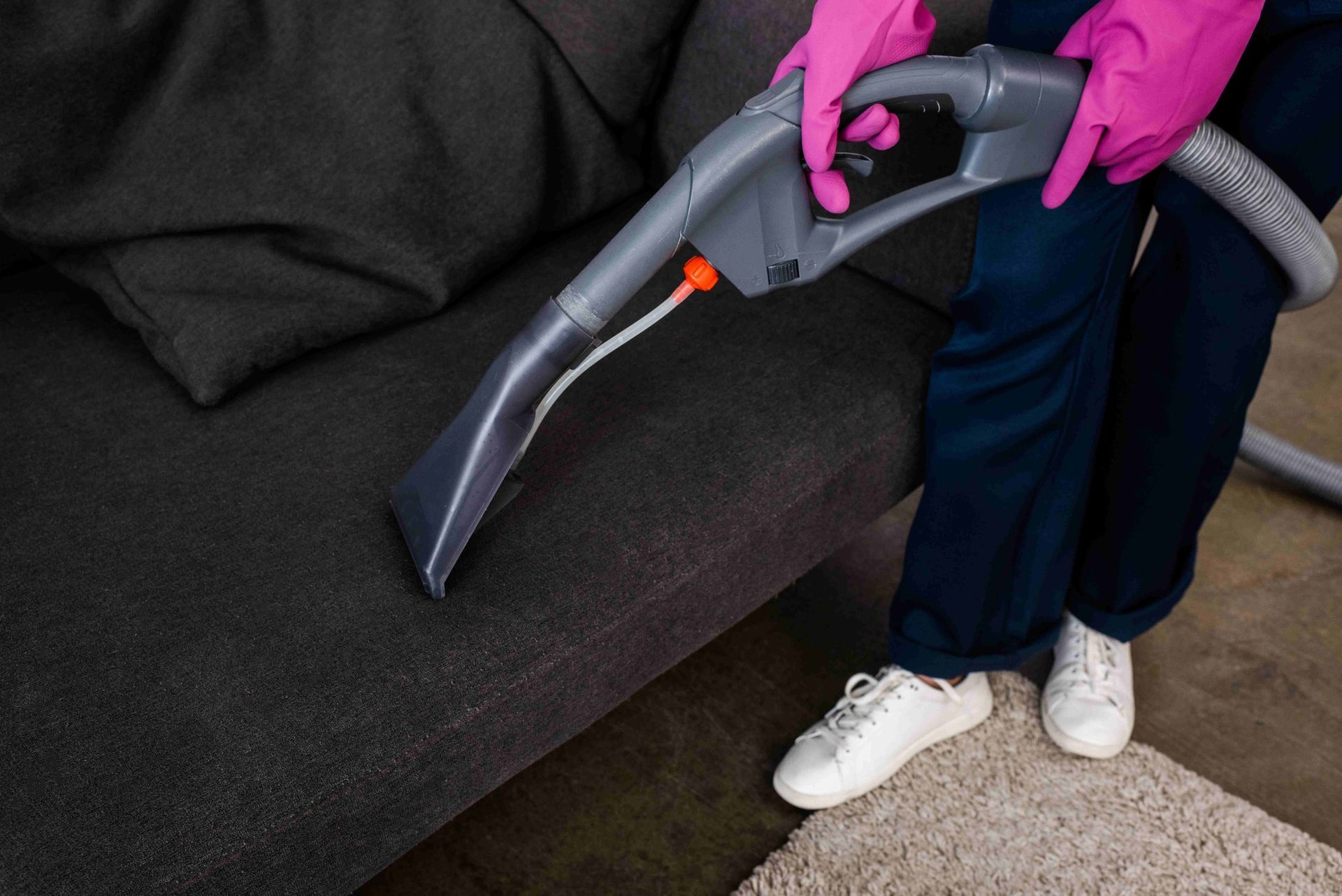 Person in pink gloves uses a vacuum hose to clean a dark sofa near a white rug.