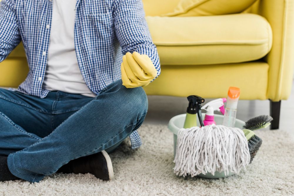 A person in a plaid shirt and yellow rubber gloves sits on a rug near a bucket of cleaning supplies and a sofa.