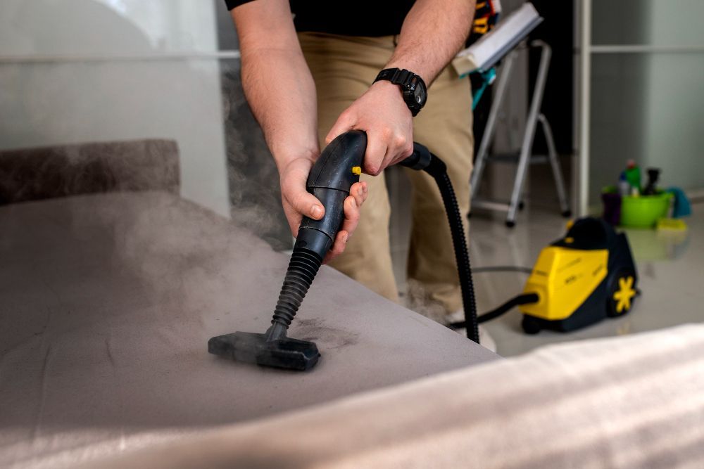 A person uses a handheld yellow and black steam cleaner to sanitize a gray sofa.