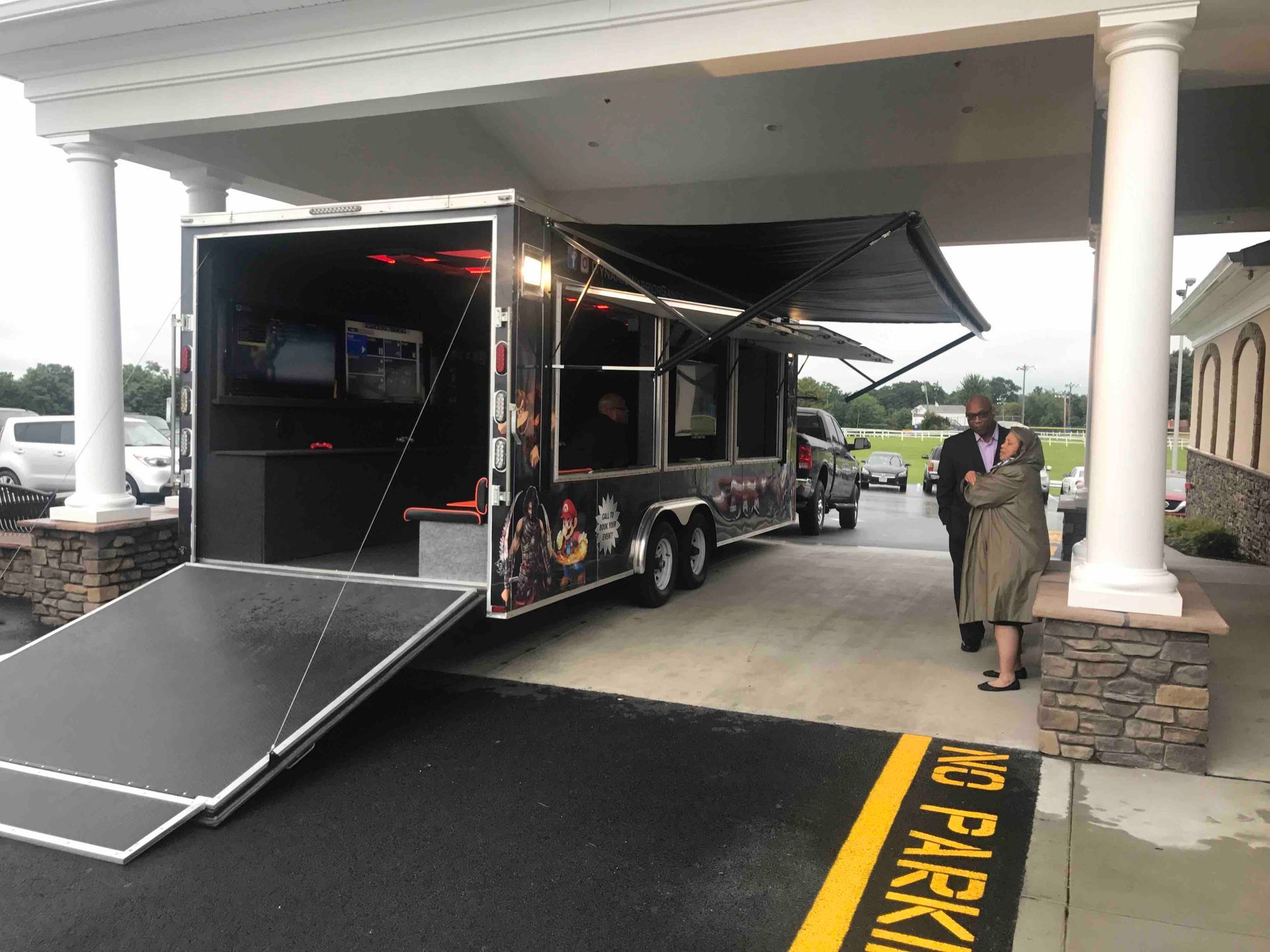 Game truck parked under an awning with ramp down. Two people walk towards the truck.