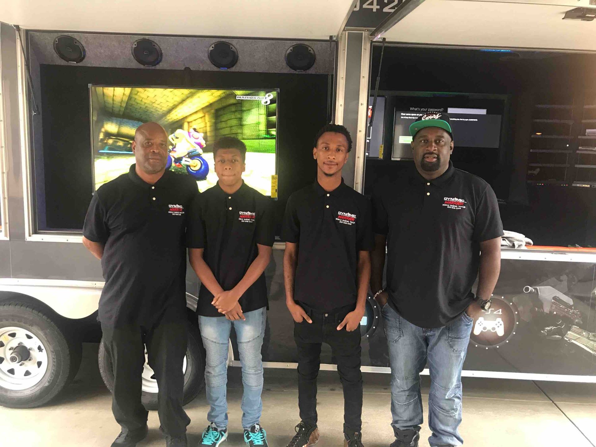 Four people stand in front of a gaming trailer, featuring a screen with a game displayed. They wear black shirts.