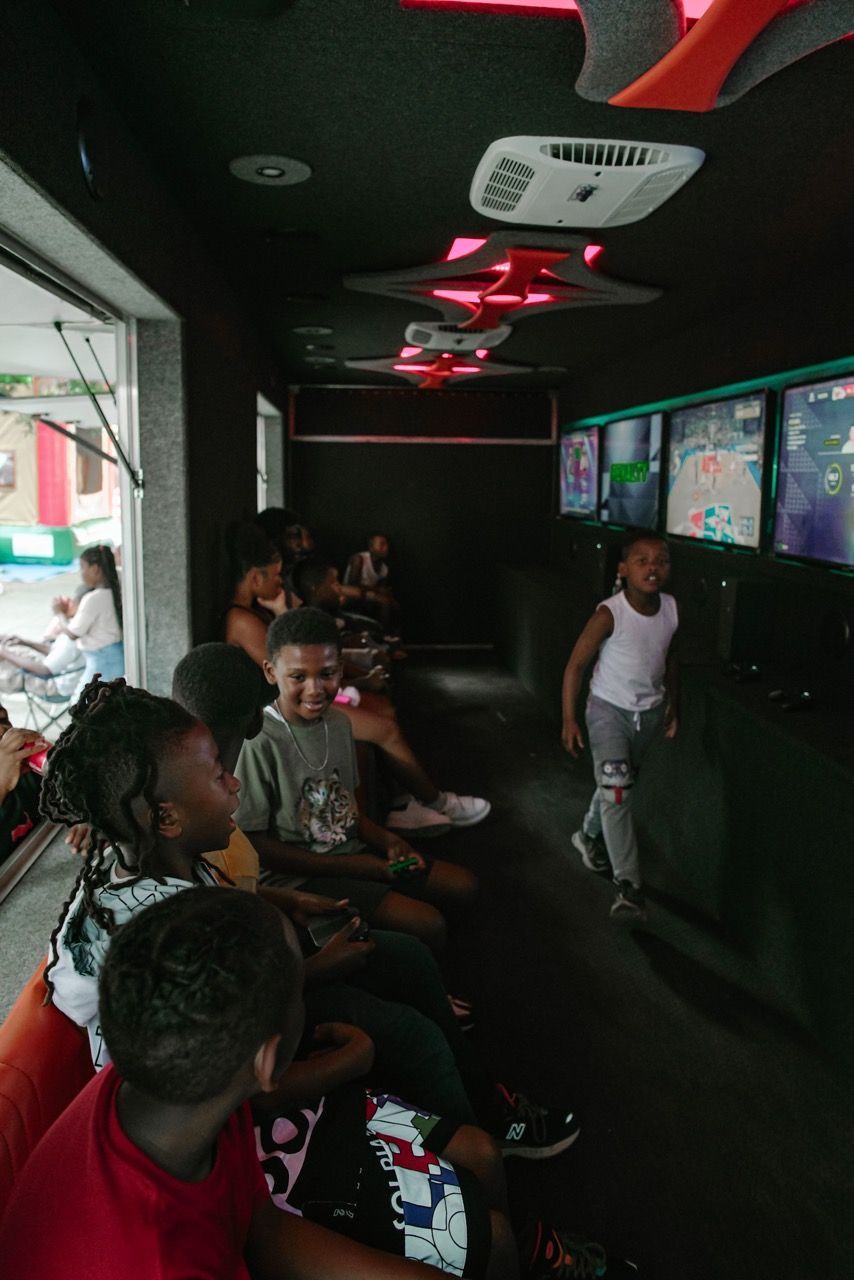 Children playing video games inside a gaming truck. Black interior, colorful lights, and monitors are visible.