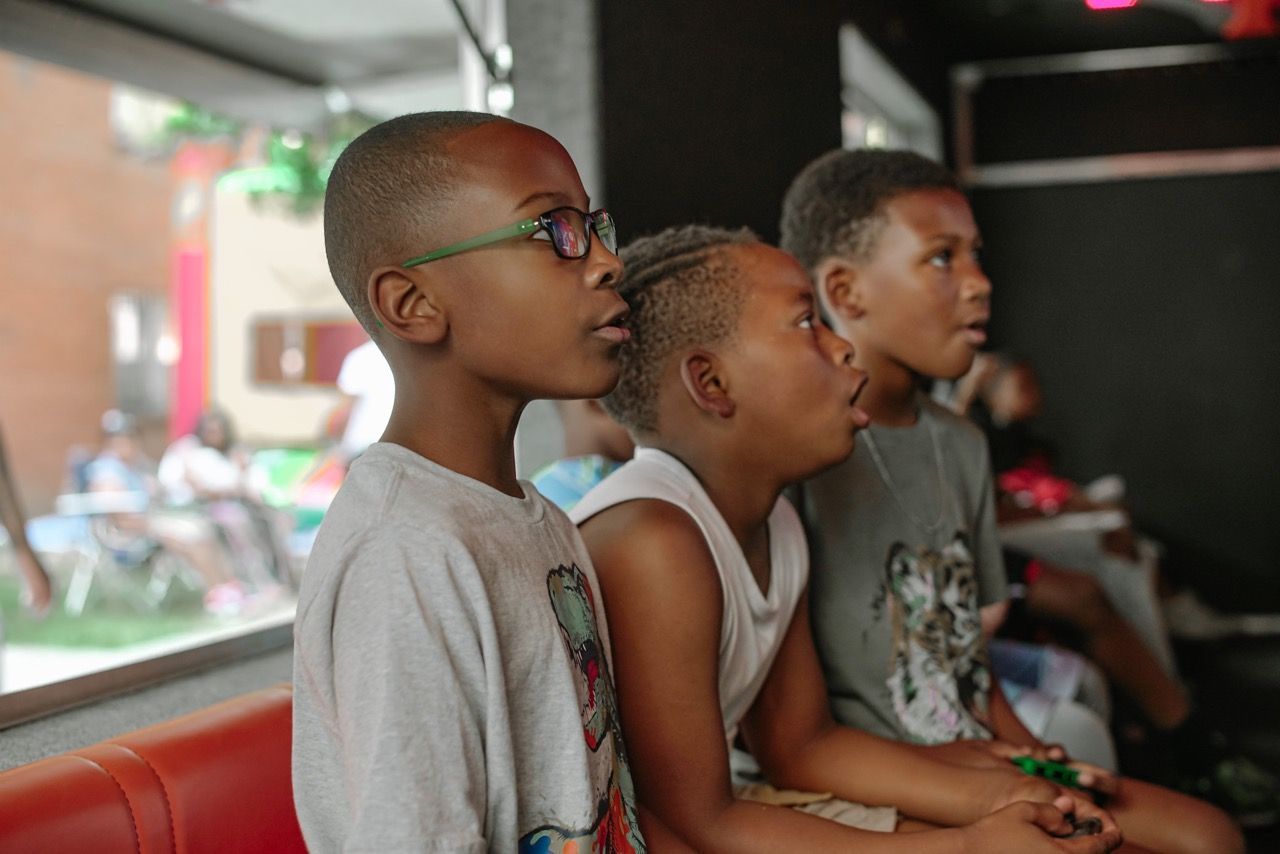 Three children watching a screen, one wearing glasses, all focused with open mouths, indoors.