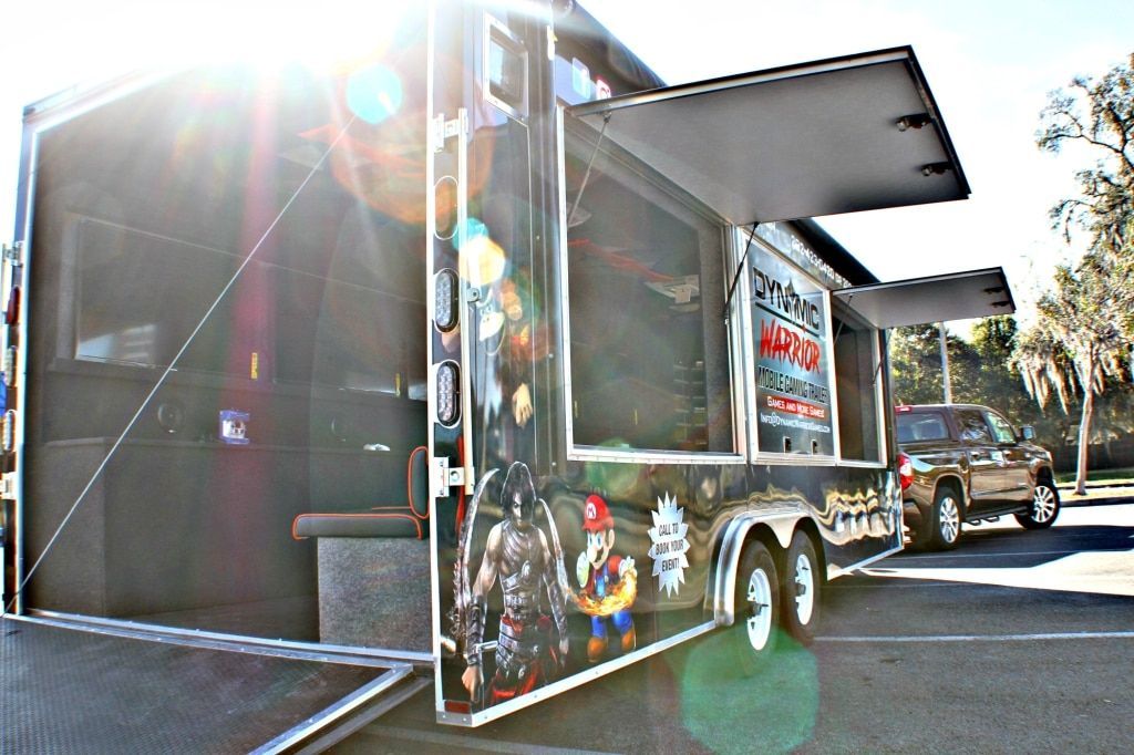 Gaming trailer with open side awning and graphics parked next to a truck in a parking lot.