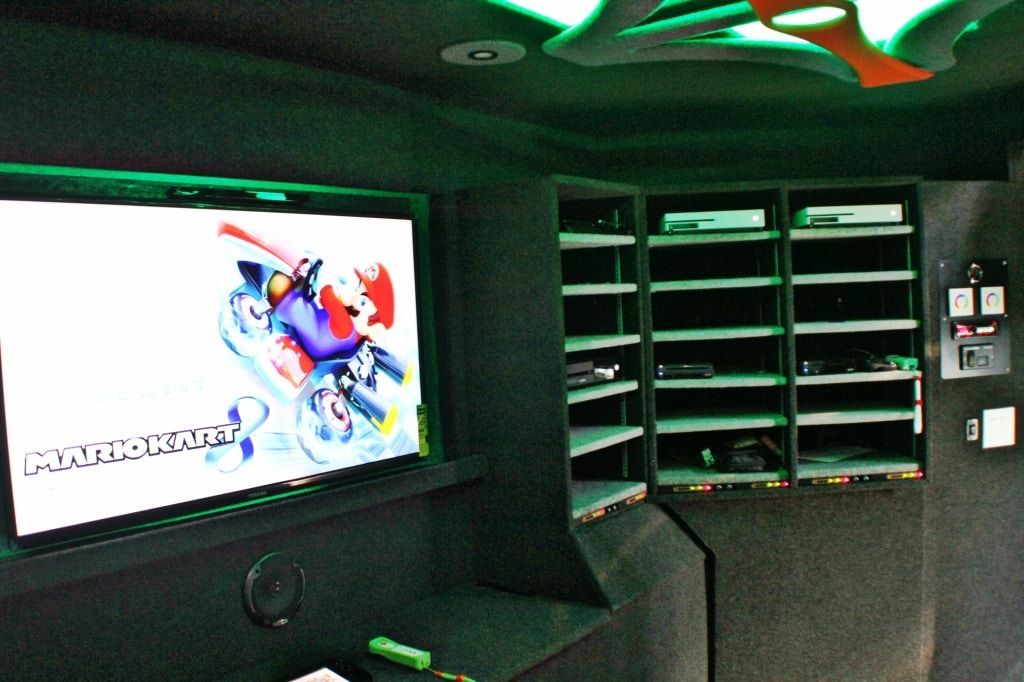 A dimly lit room with a large screen displaying Mario Kart, shelves for consoles, and green accent lighting.