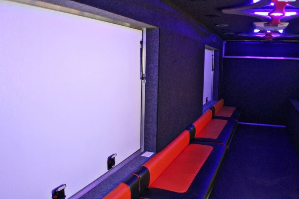 Interior of a vehicle, featuring a white screen, red and black seating, and a door; lit by blue and red lights.