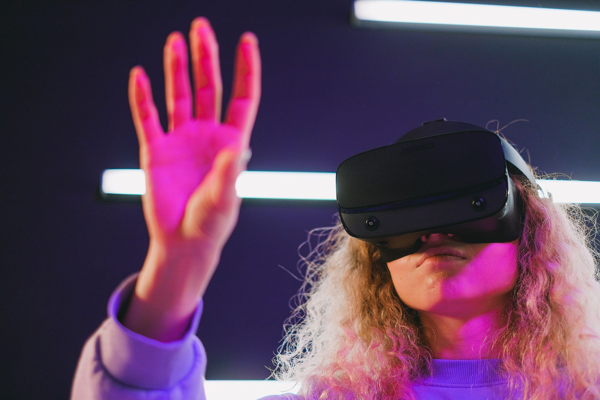 Person wearing a VR headset, with a hand raised in front of them, lit by pink and purple light.