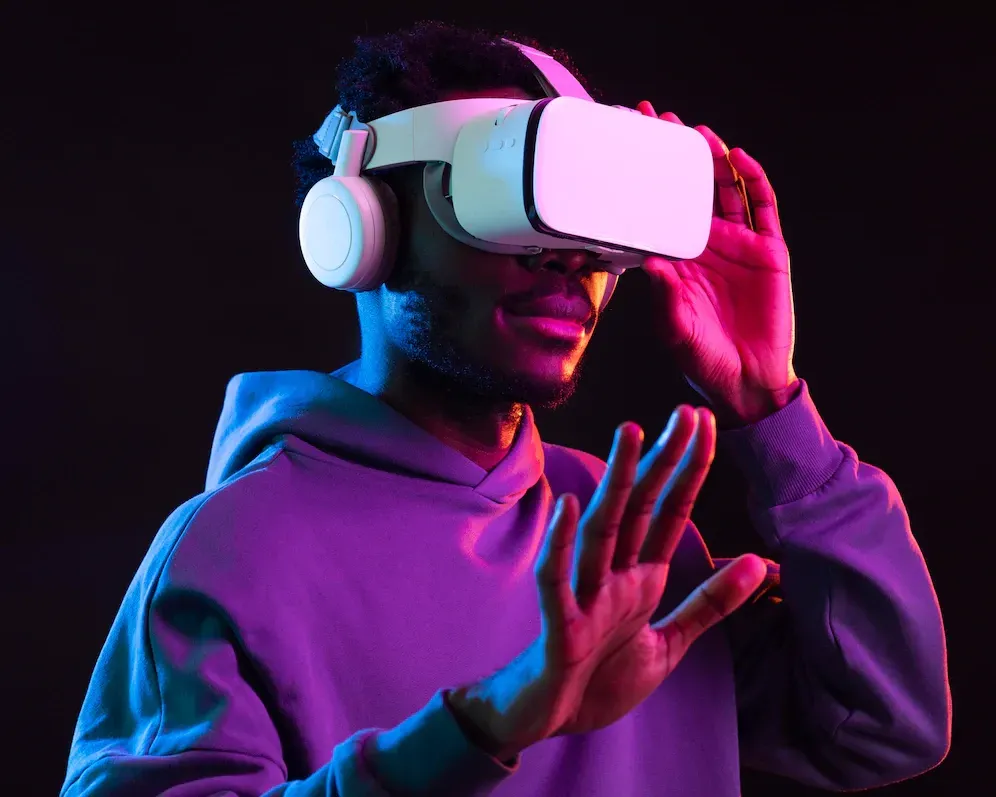 Person wearing VR headset, hands up, illuminated by purple and pink lights against a dark background.