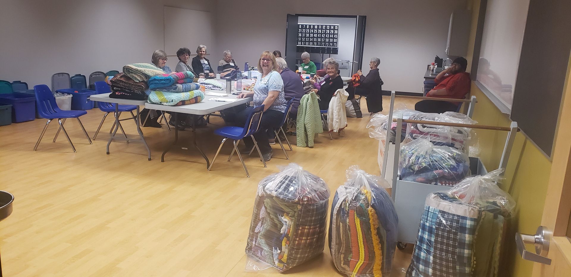 A group of people are sitting at tables in a room with bags of clothes.
