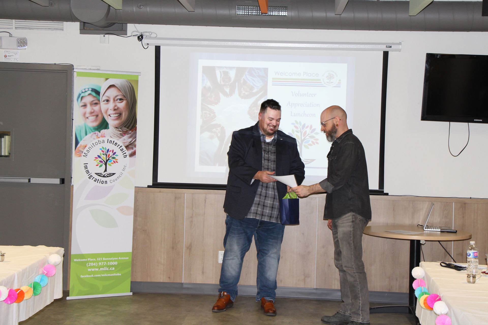 Two men are shaking hands in front of a projector screen in a room.