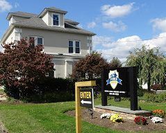 Exterior of beige building with sign for a business. Enter sign in foreground. Flowers and shrubs are in the yard.