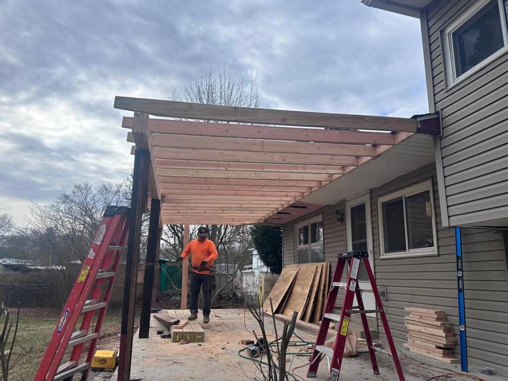 Man building a wooden pergola attached to a house. Cloudy sky, ladders, and building materials visible.