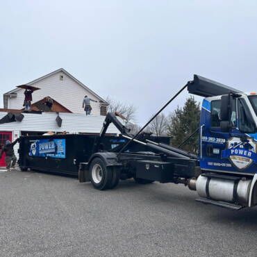 Workers removing roof shingles, loading debris into dumpster, beside white building. Blue Power Truck present.