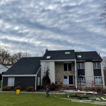 House with dark gray roof, siding, and white trim. Man walking on lawn under cloudy sky.