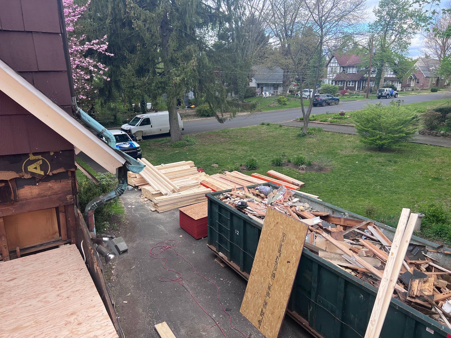 Construction site with debris container; lumber; blue and white truck; houses and trees in the background.