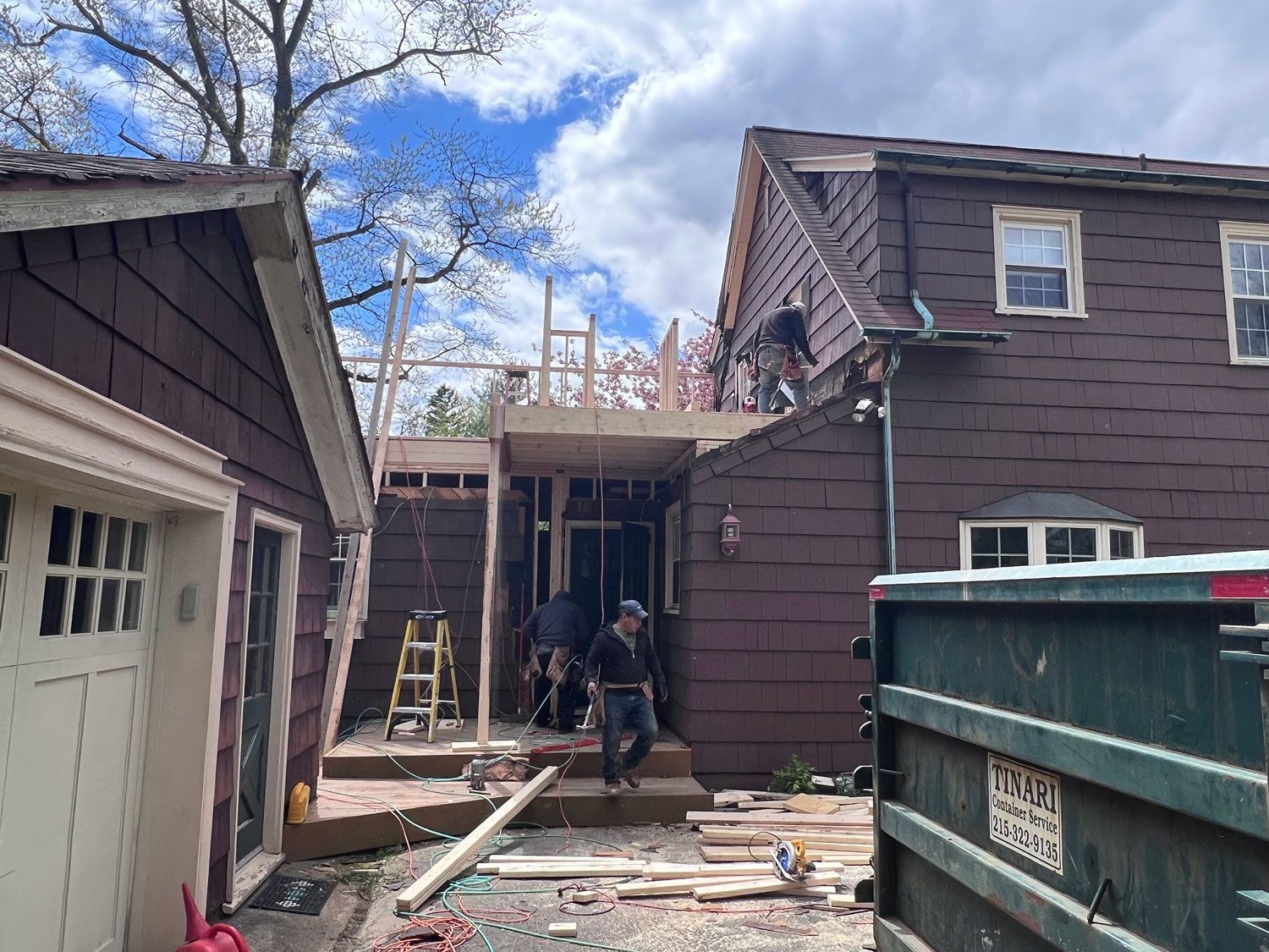 Construction site: workers building a house addition. Brown siding, blue sky, lumber, and a dumpster are visible.