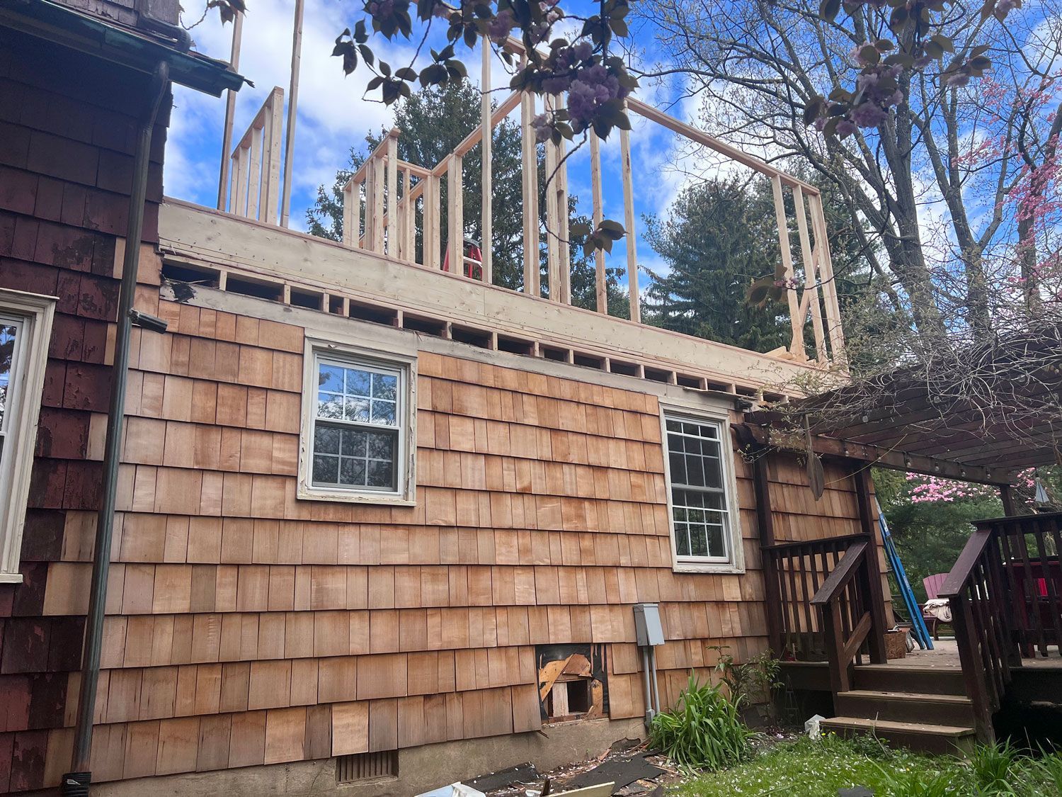 House under construction, wood-shingled exterior, framing for second story addition, blue sky.
