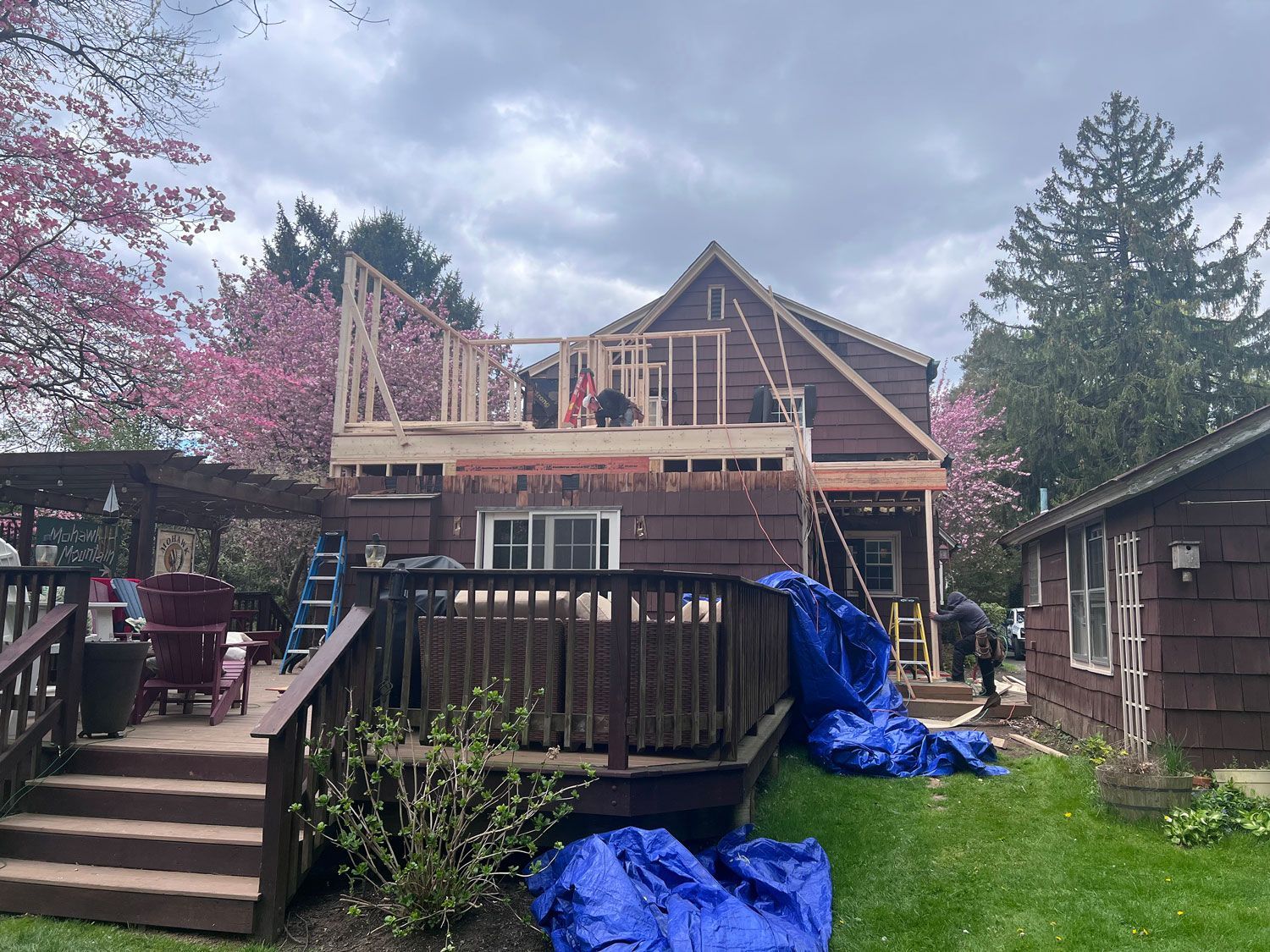House under construction: wooden frame added above the existing roof. Workers on the roof. Cloudy day.