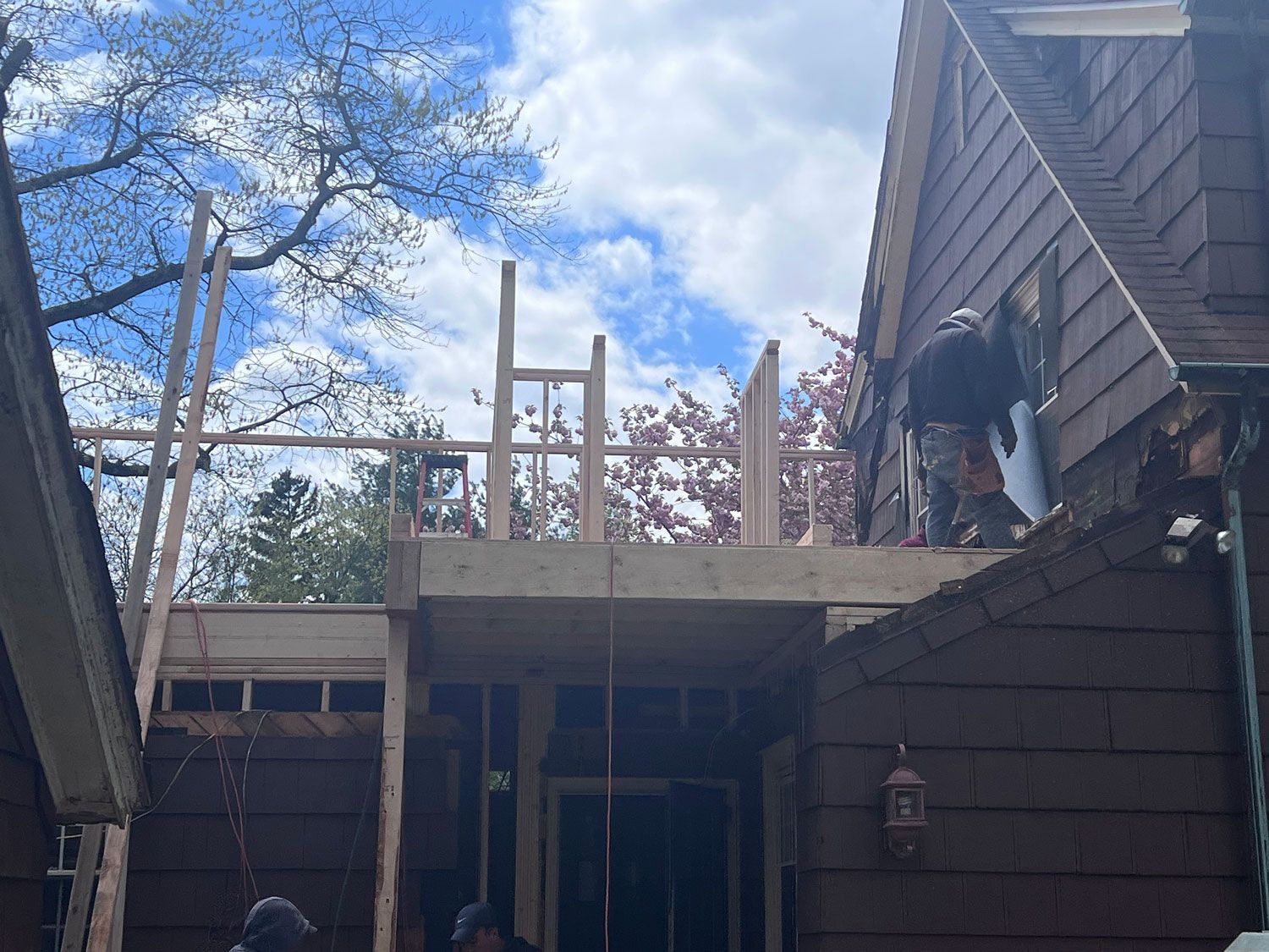 Construction workers building a wooden deck addition on a brown shingled house on a sunny day.
