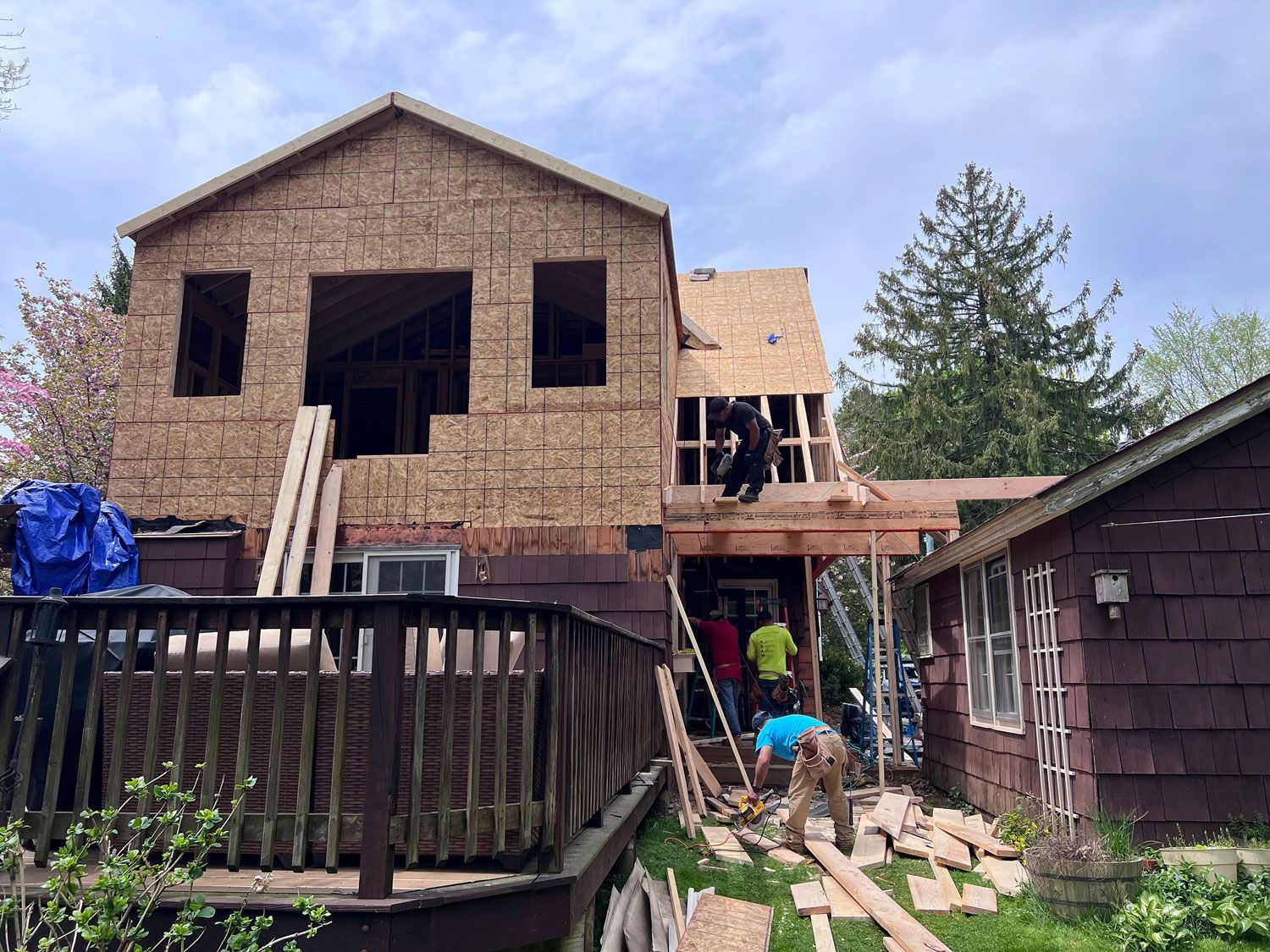 Construction workers building a two-story addition to a house. Workers on a deck, roof, and ground. Cloudy day.