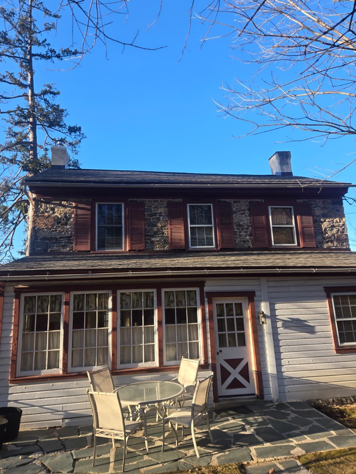Two-story rustic house with red shutters and patio chairs in front under a clear blue sky