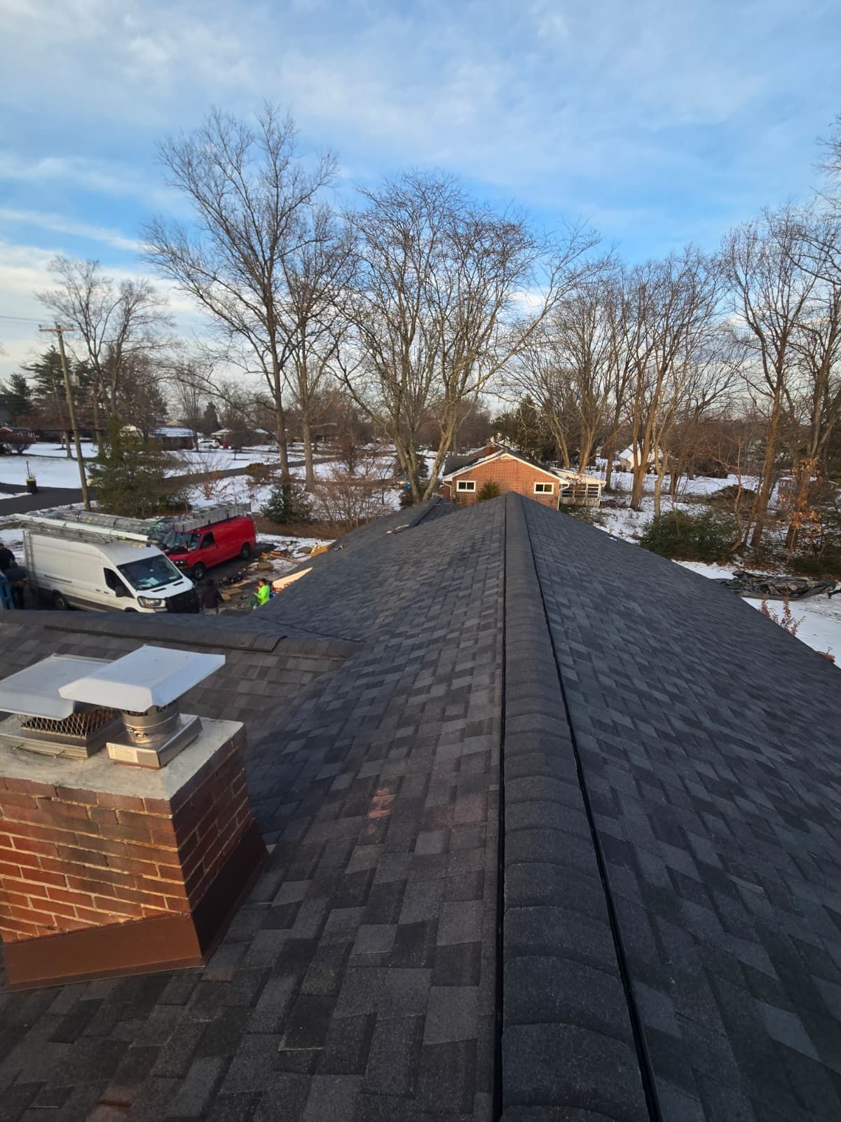 View along a shingled roof toward a snowy yard with bare trees and a blue sky