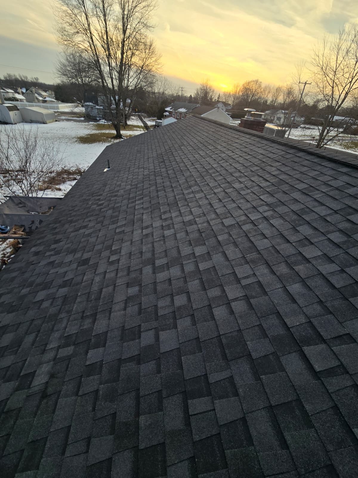 A close-up view of dark grey asphalt shingles on a roof at sunset, with a snowy landscape visible in the background.