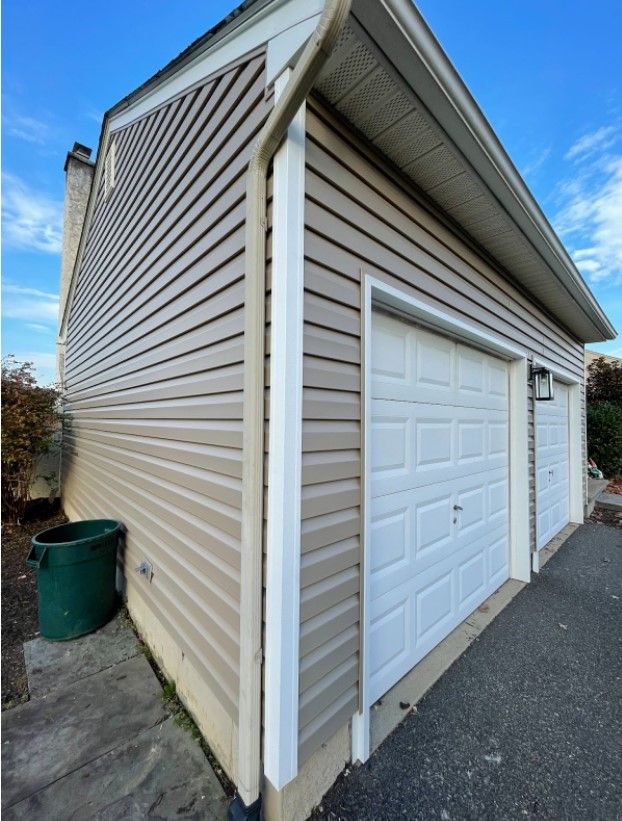 Tan-sided garage with white doors and trim, green trash can, blue sky.