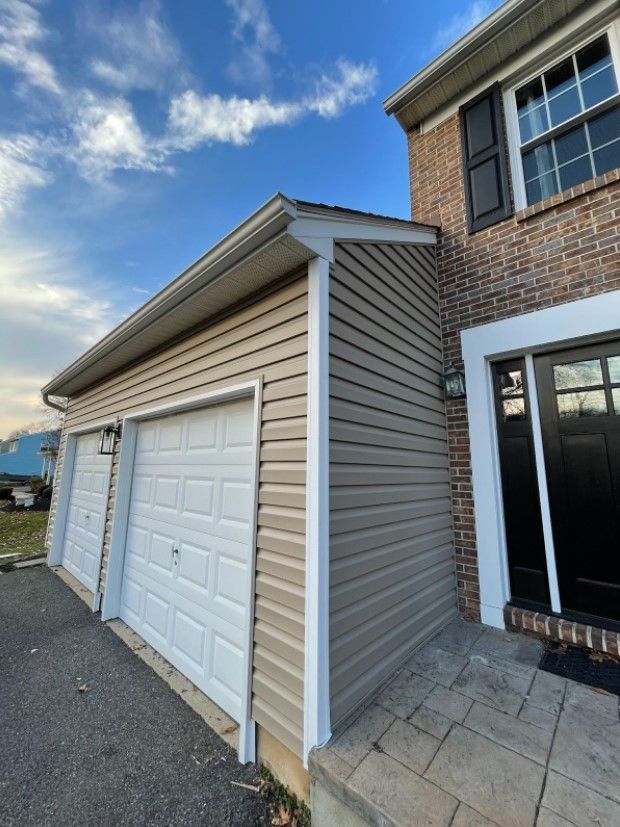 Tan vinyl-sided garage next to a brick house. White garage doors and trim. Cloudy blue sky.