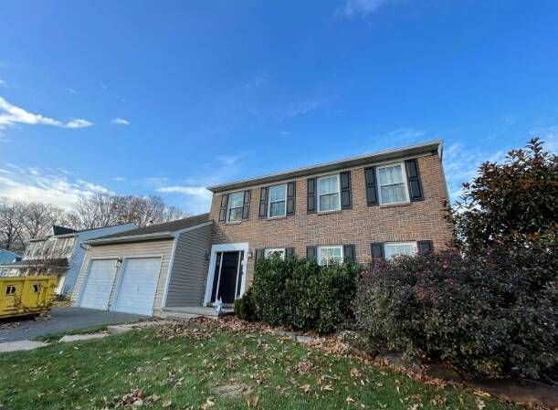 Two-story brick home with gray siding, black shutters, blue sky. Two-car garage, front bushes, and fallen leaves.