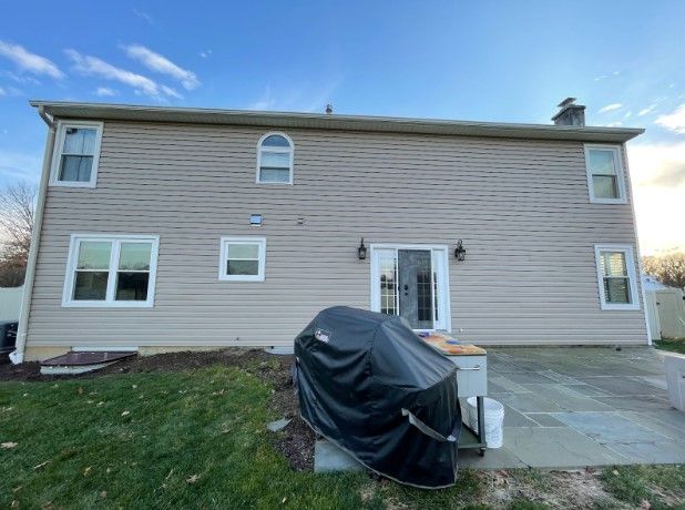 Back of a two-story beige house with a grill on a patio and a grassy yard under a partly cloudy sky.