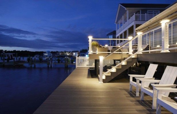 Deck with white chairs, steps, and lighting at night overlooking a waterfront.