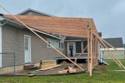 House with porch under construction; wooden frame and roof partially built, beige siding, cloudy day.