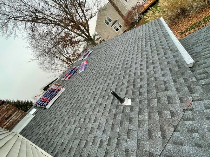 Gray shingle roof with a vent pipe and stacks of shingles. A house and trees are in the background.