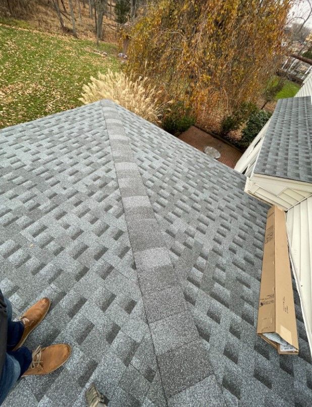 Gray shingle roof with ridge line, with a brown box of shingles. A person stands on the roof.