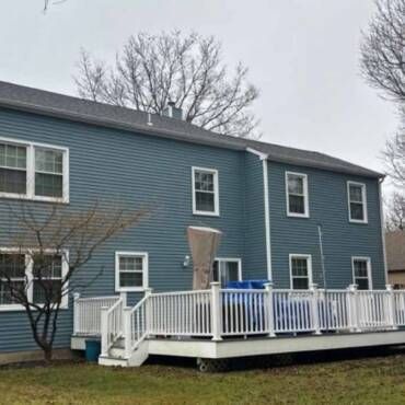 Back of a two-story blue house with a white deck, blue trash cans, and bare trees under a gray sky.