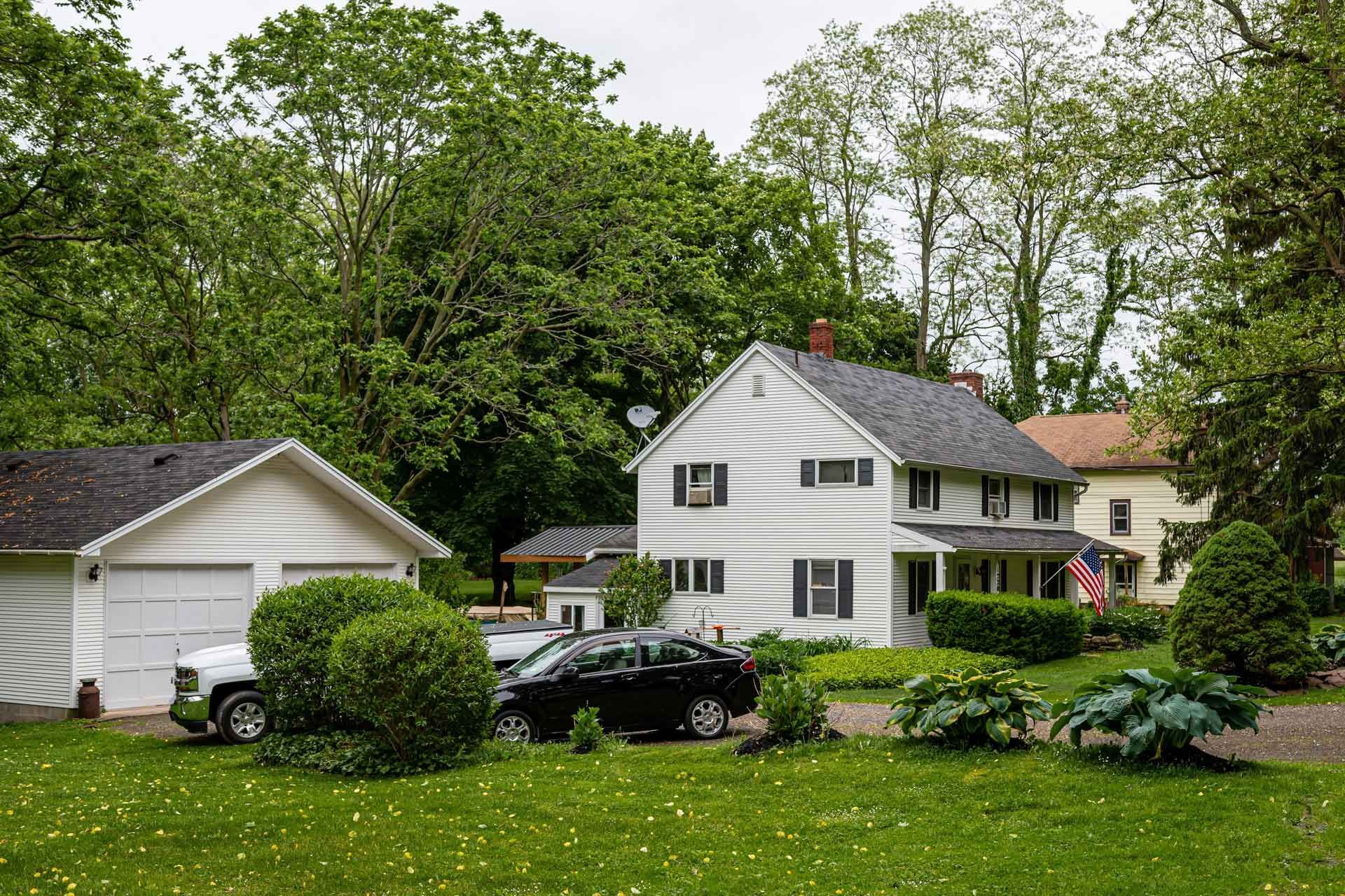 White house with attached garage and cars parked in front, set in a green yard with trees.