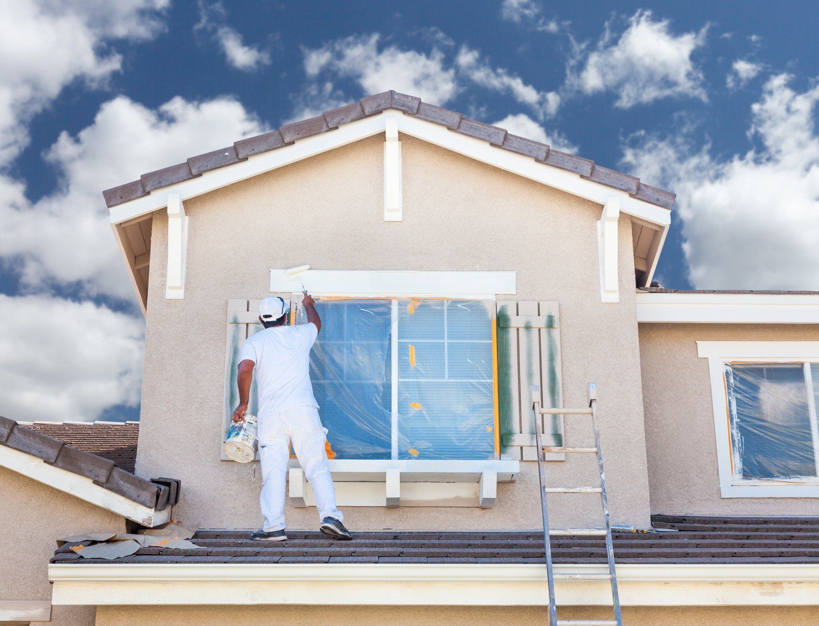 Painter on ladder, painting window trim on beige house. Blue sky with clouds.