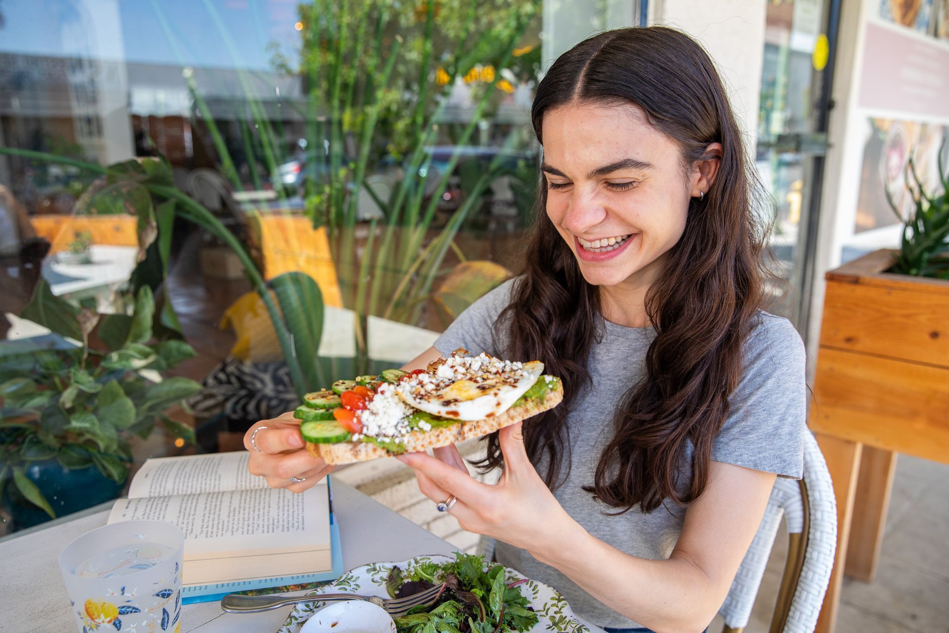 Woman smiling, holding avocado toast with egg and veggies, seated outside.