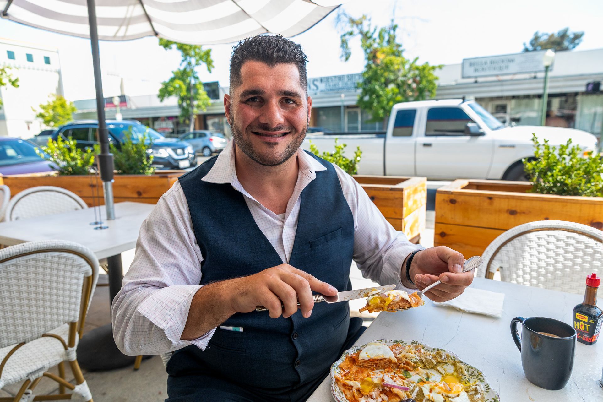 Man in vest seated at outdoor table, eating. Sunny day, white table, buildings in background.