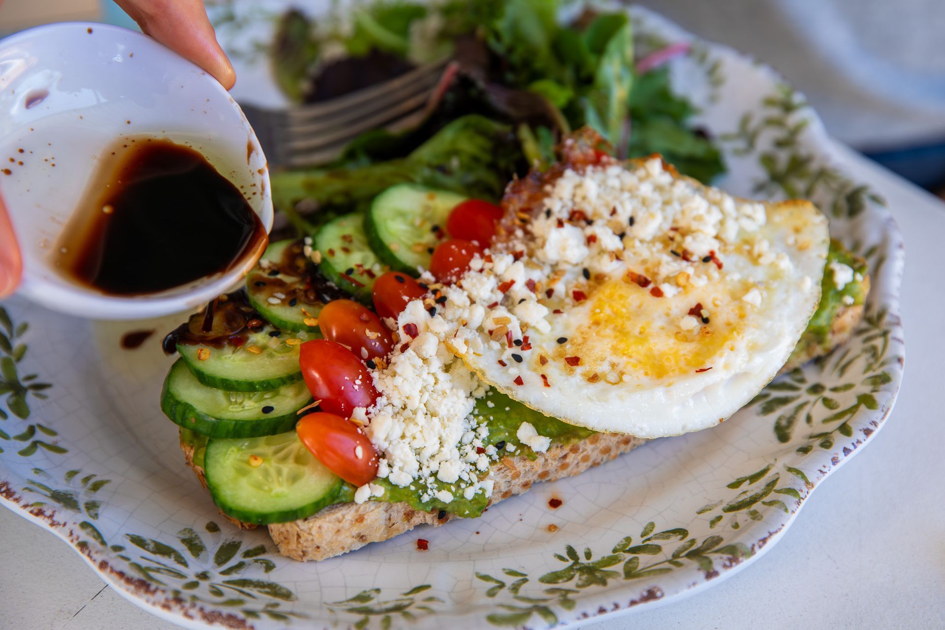 Avocado toast topped with egg, cucumber, tomatoes, and feta, being drizzled with balsamic vinaigrette.