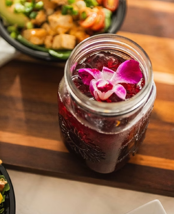 Jar of red drink with pink flower, on a wooden board next to a salad.