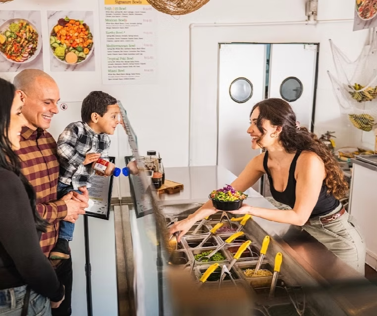 Three people smelling food bowls indoors; neon sign says