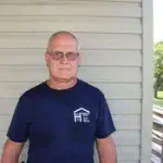 A man wearing glasses and a blue shirt is standing in front of a white wall.